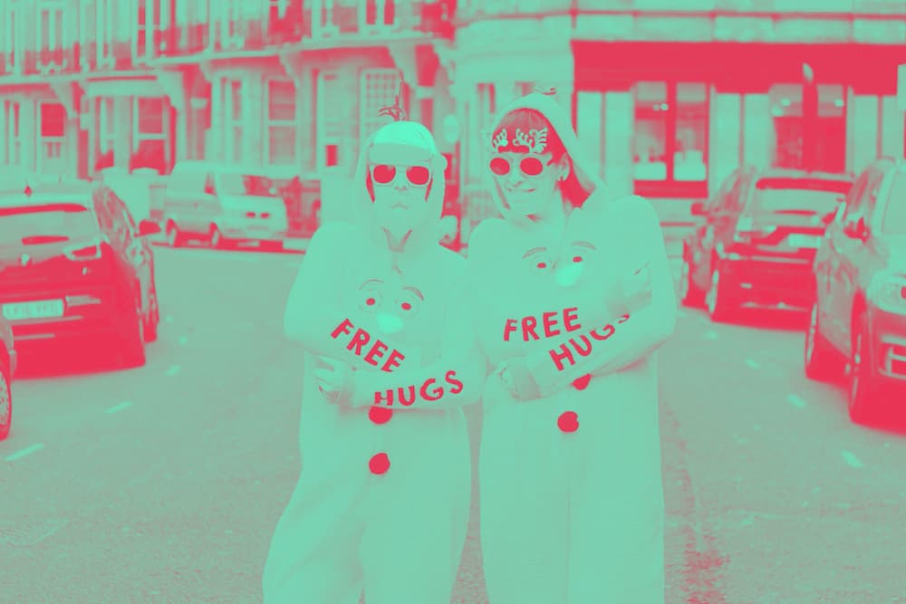 A man and a woman in a shopping street, dressed in Christmas onesies that read "free hugs" on their sleeves.