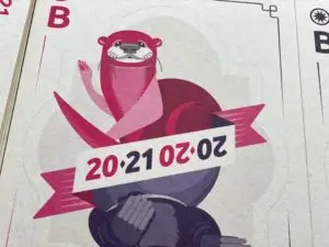 Der Fischotter-Teil der Postkarte, die wie eine Spielkarte gestaltet ist. Man sieht oben einen pinkfarbenen Fischotter. Darunter ein Band, auf dem die Jahreszahlen "2020" und "2021" diagonal dargestellt sind.