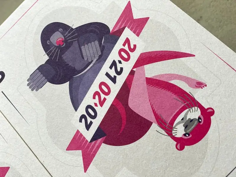 Postkarte, die wie eine Spielkarte gestaltet ist. Man sieht links oben einen violetten Maulwurf, rechts unten einen pinkfarbenen Fischotter. Dazwischen ein Band, auf dem die Jahreszahlen "2020" und "2021" diagonal dargestellt sind.