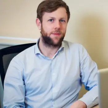 Chris Stainthorpe looking up from his laptop with an attentive expression. He’s wearing a casual beard, short hair and a light blue shirt with the top buttons open.