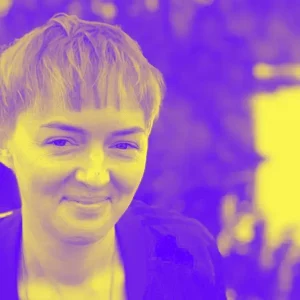 Yellow-purple duotone image of Sabine Harnau. She’s wearing short hair, a training jacket and a slightly mischievous smile.