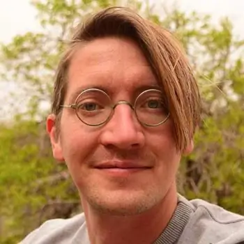 Gerrit Schuster ist in der Natur und lächelt selbstbewusst. Er trägt halblanges Haar mit Seitenscheitel, eine John-Lennon-artige Metallbrille, Dreitagebart und ein graues T-Shirt.
