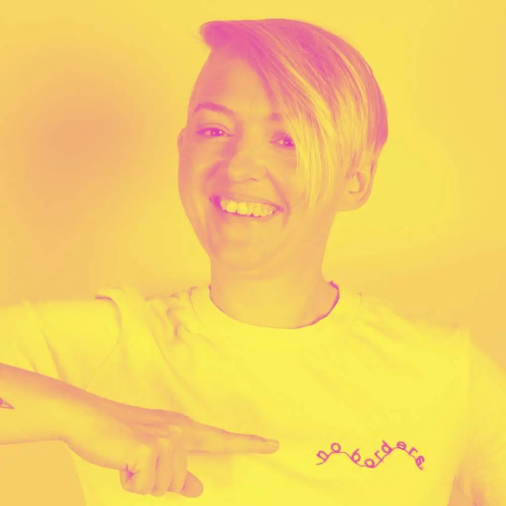Yellow-pink duotone image of Sabine Harnau smiling and pointing at the embroidery on her T-shirt. It reads “no borders”.