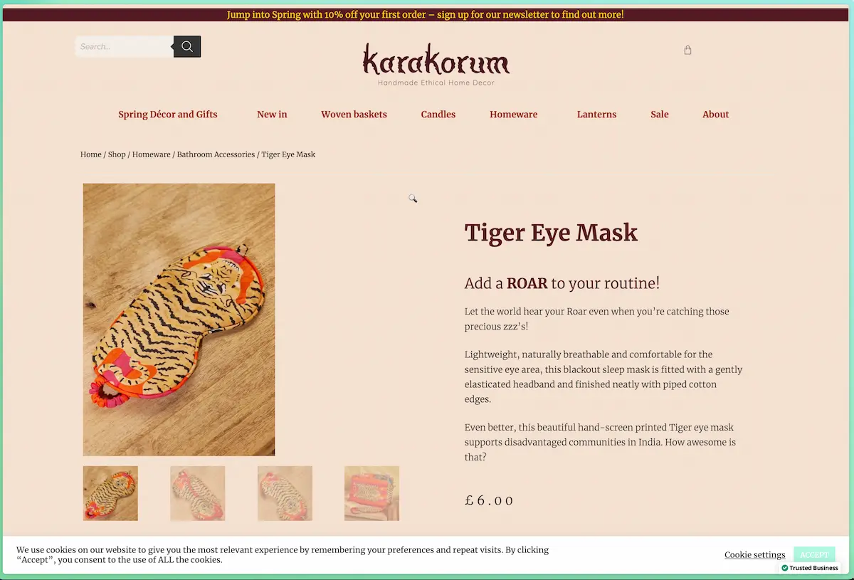Product detail page for the Tiger Eye Mask at Karakorum. The background is a strong beige, with text in dark red and brown. The product is beige with a red and brown pattern. It is photographed on a wooden background that is also beige with a brown pattern. At the bottom of the screenshot, there is a white cookie notice with a text link leading to “cookie settings” and a green button saying “accept”.
