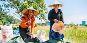 2 indonesische Reisbäuer*innen in traditioneller Kleidung lächeln dich an. Der Mann links zeigt den Daumen nach oben, die Frau rechts hält einen Korb. Im Hintergrund sind Bäume, Felder und Silos zu sehen.