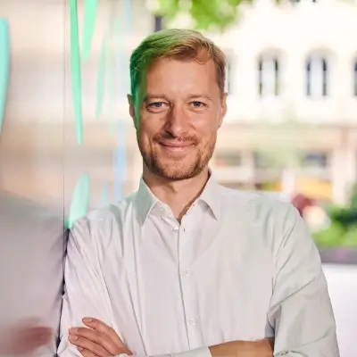 Professor Fabian Diefenbach smiles at you. He's got his arms crossed in front of his chest and leans against a wall to his right. He has short reddish hair, a slight beard and wears a white shirt. The background is blurred.
