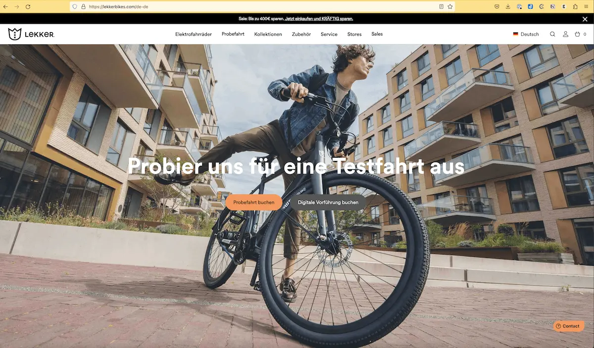 Screenshot der Lekkerbikes-Startseite. Ein junger hipper Typ steigt in einer Neubausiedlung mit Apartments auf sein Lekkerbike. Dabei siehst du ihn von links unten. Er schaut von dir weg. Überschrift: Probier uns für eine Testfahrt aus, Button links, orange: Probefahrt buchen, grauer Button rechts: Digitale Vorführung buchen