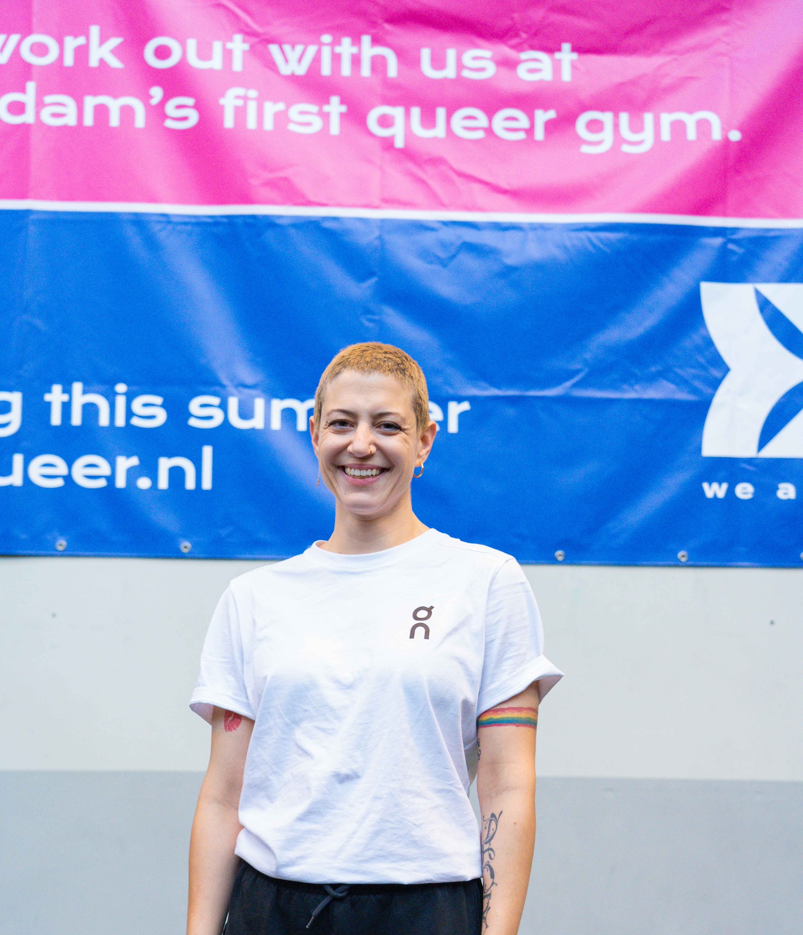 Ein Mitglied des We Are Queer Trainingsteam strahlt dich an. Die Person trägt sehr kurze Haare, große Creolen-Ohrringe, ein T-Shirt von On und ein leuchtendes Regenbogen-Tattoo am Oberarm. Im Hintergrund hängen Poster von We Are Queer, die erklären, dass dies das erste queere Gym von Amsterdam ist.