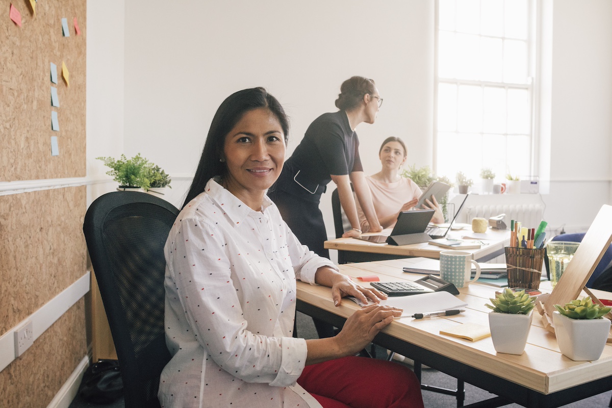 A female employee is sitting at her desk. She has turned to look at you. There are pens, a calculator, a mug and plants on her desk. Behind her, post-it notes are attached to a large cork board. In the background, 2 of her colleagues are talking at another desk.