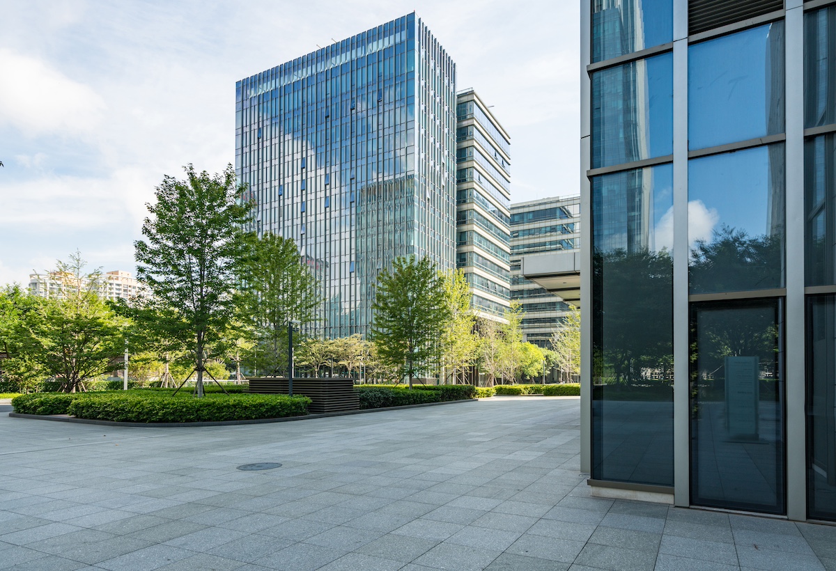 Modern glass-and-steel office buildings in a landscaped business park. There are trees, hedges, green areas and paved areas.
