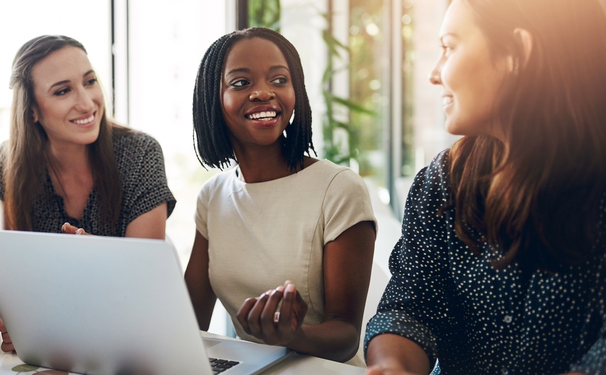 3 business women are talking and smiling while looking at a laptop.