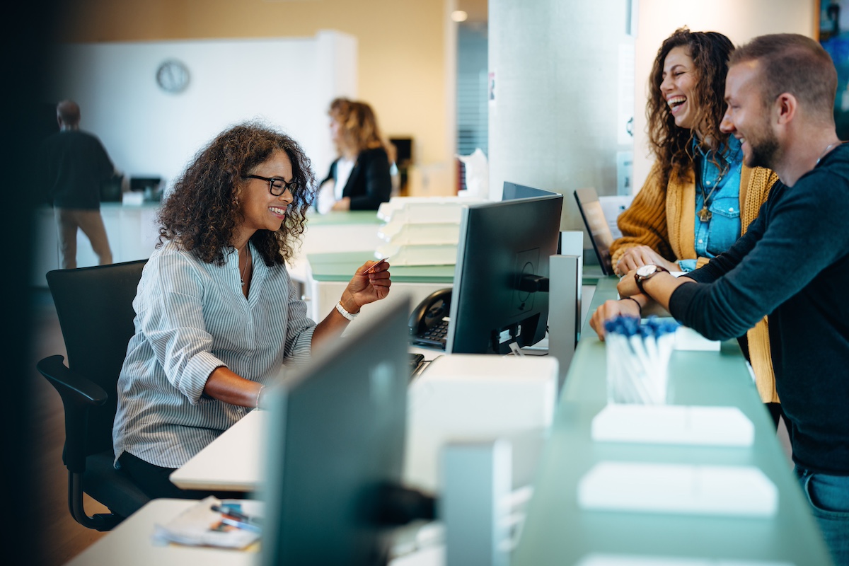 People standing at a municipal office reception and talking to a receptionist. The receptionist is smiling and working at the computer. The people waiting at the front desk are smiling and laughing.