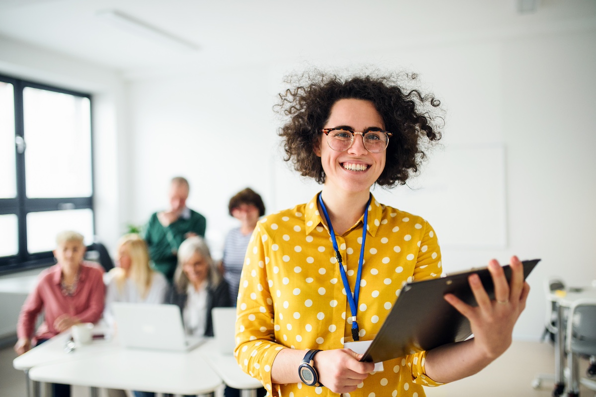 A woman holds a tablet while a group looks at computers behind her. She is teaching a workshop.