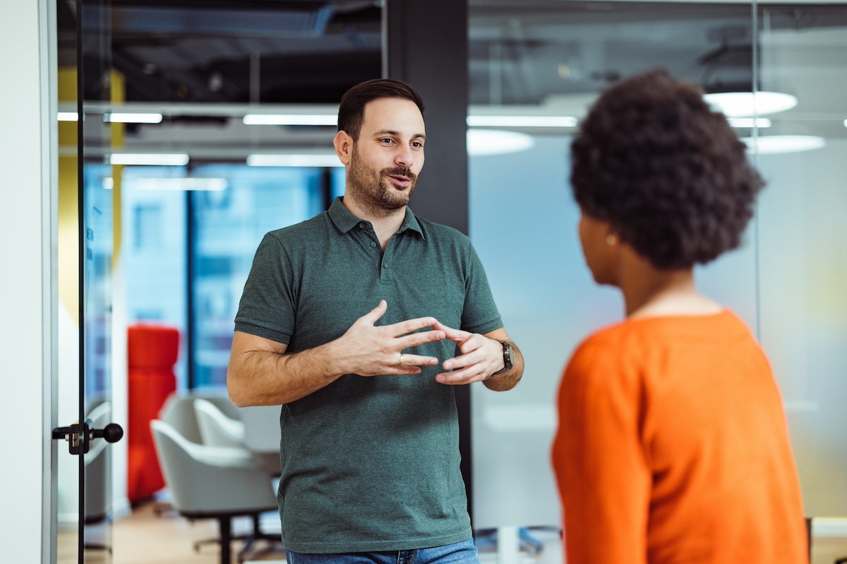 A smiling man is talking to a woman who is looking at him, with her back turned to you. There's a meeting room in the background.