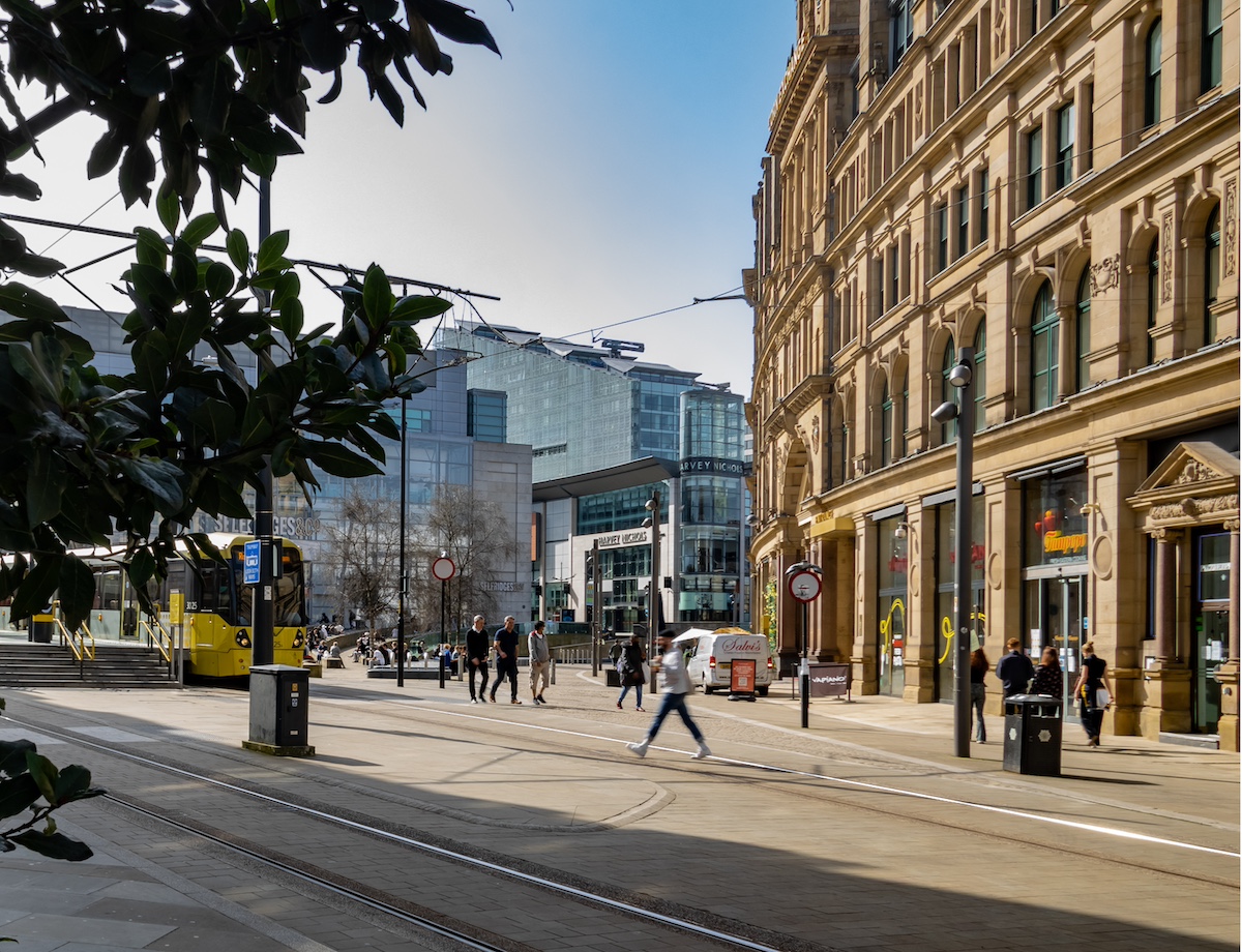 Manchester City centre in the UK. There are different buildings, old and new. People are walking and crossing the street. A yellow tram is in the background, and a tree with shade is in the foreground.