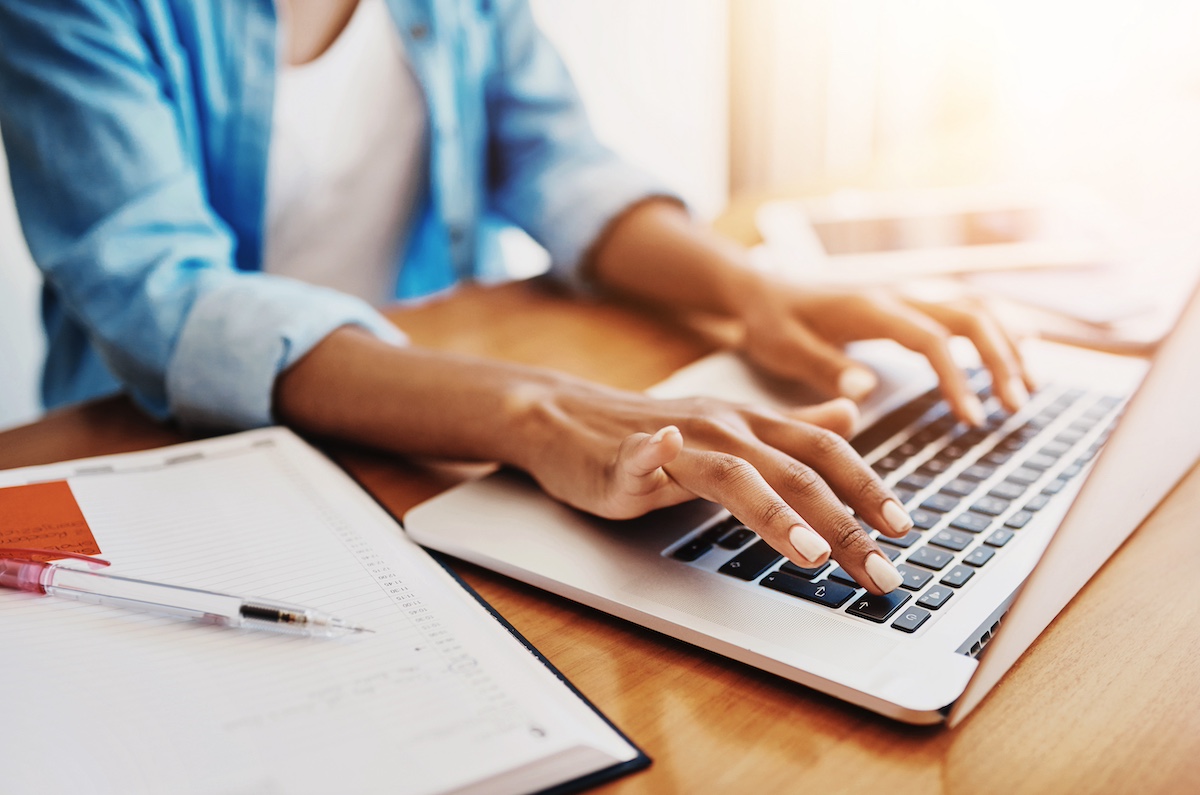 A woman is typing at a laptop. She has a note pad and pen to her right. Only her arms and hands are shown.